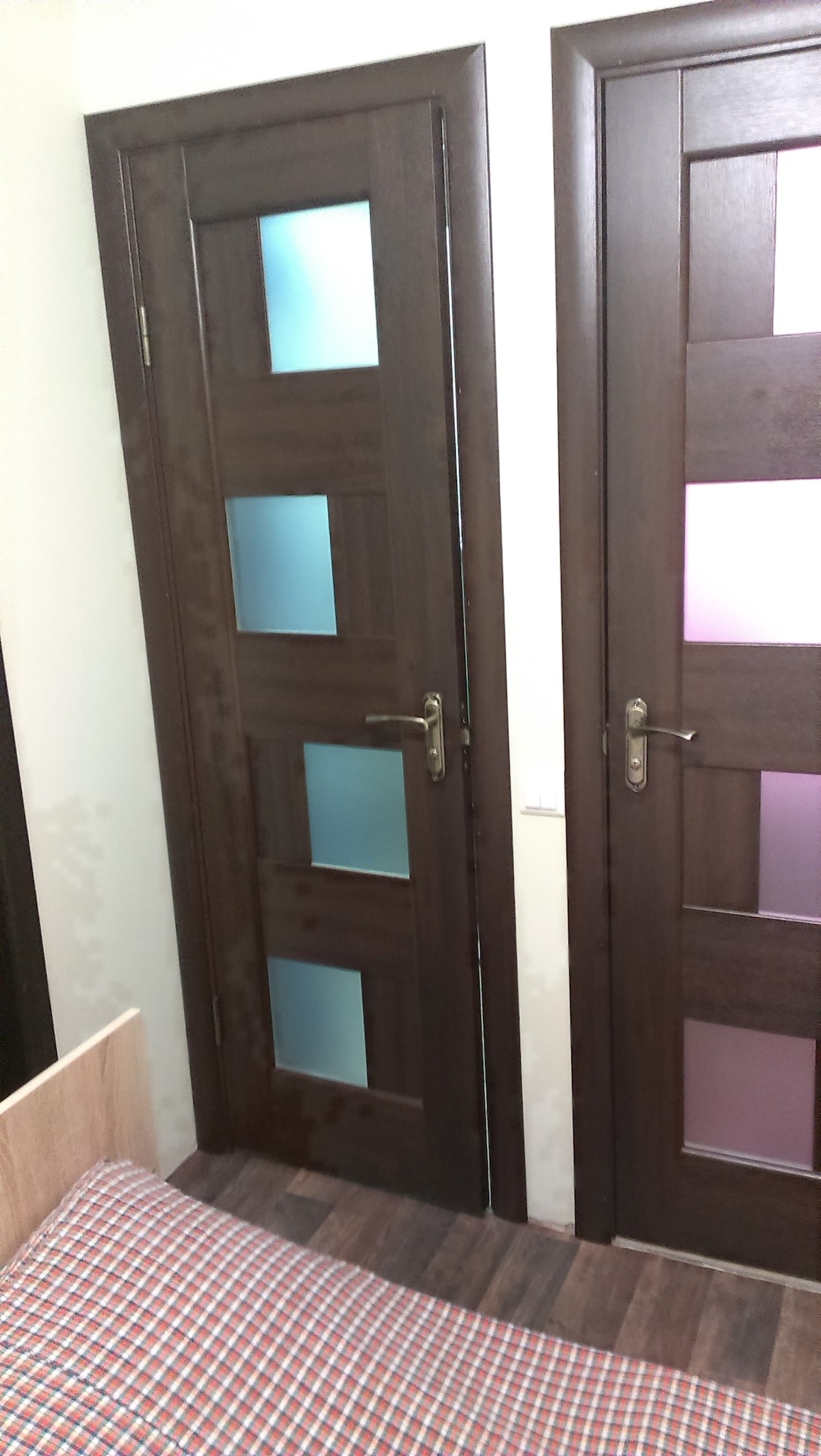 Two wooden doors with glass panels are displayed, featuring blue and pink glass inserts that provide a hint of color. The wood finish contrasts with the light-toned wall. A portion of a wooden bed frame is visible in the foreground on a checkered patterned bedspread.