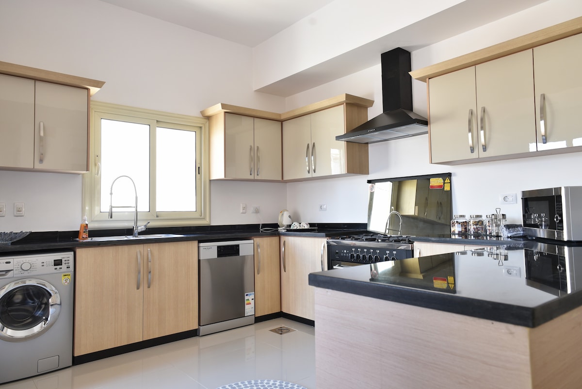 A spacious kitchen features modern cabinetry in light wood and sleek black countertops. New appliances are visible, including a dishwasher, gas stove, and washing machine. Natural light filters through a window, illuminating the kitchen's open layout and functional design.
