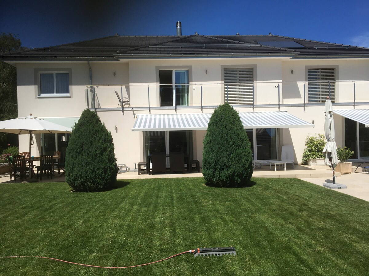 A spacious outdoor area is depicted, featuring a well-maintained lawn and manicured shrubbery. Two large evergreen trees frame the patio, which is equipped with a dining set under an awning. A second patio umbrella is positioned for shade, contributing to a relaxing atmosphere.