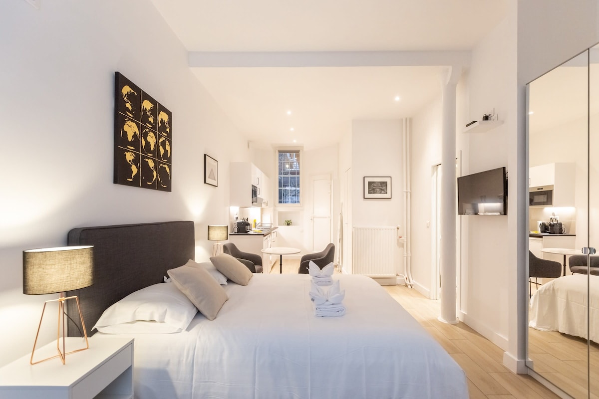 A modern studio features a spacious bed with neutral bedding and scattered decorative pillows. Soft lighting is provided by bedside lamps. An open kitchen area is visible in the background, along with a dining table and chairs. A full-length mirror adds to the sense of space.