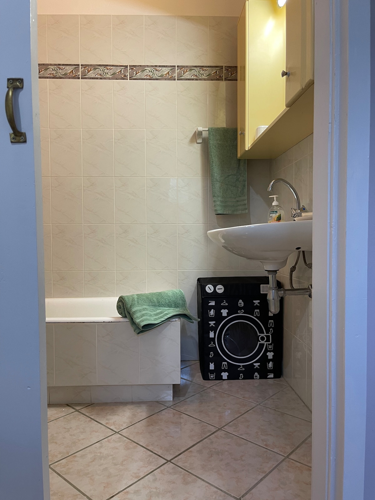 A compact bathroom features light tile flooring and beige wall tiles. A white bathtub rests against one wall, accompanied by a green towel placed neatly on the edge. Above the sink, a cabinet with yellow doors offers storage, while a black laundry basket with a patterned design sits nearby.