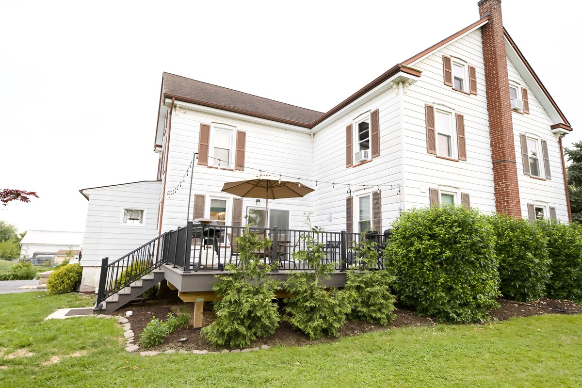 The exterior of a two-story house features white siding and brown shutters. A spacious deck extends from the kitchen, furnished with a table and chairs. Lush green bushes surround the deck, enhancing the outdoor space. A well-kept lawn leads to the home's entrance.