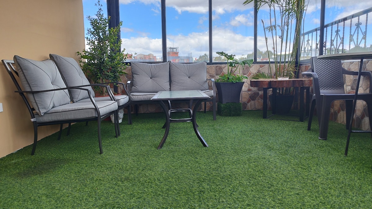 A shaded terrace features comfortable seating with gray cushioned chairs and a glass-top table. Lush green artificial grass provides a soft surface, while potted plants add a touch of greenery against the backdrop of a clear blue sky.