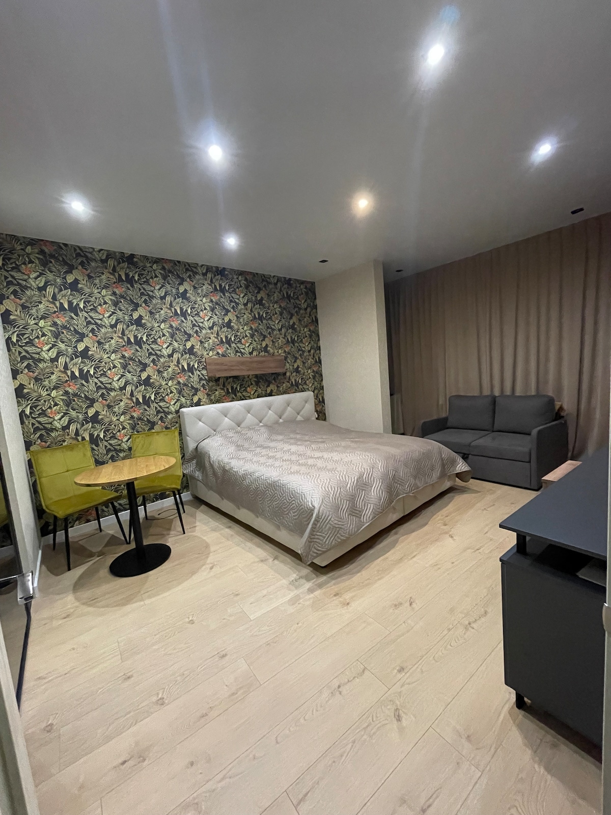 A modern bedroom space features a king-size bed with a light-colored quilt and a patterned accent wall of greenery. A small round table with two chairs is positioned near the bed, and a gray sofa is situated against the wall, contributing to the overall comfort of the room.