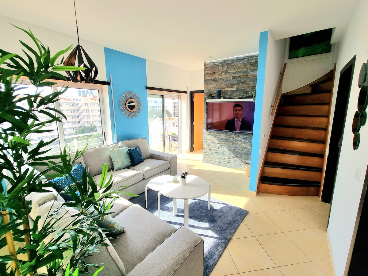 A modern living area is featured, showcasing a comfortable sectional sofa and a round coffee table on a soft area rug. Large windows allow ample natural light, and a staircase is visible against a blue accent wall. A television is mounted on a stone feature wall.