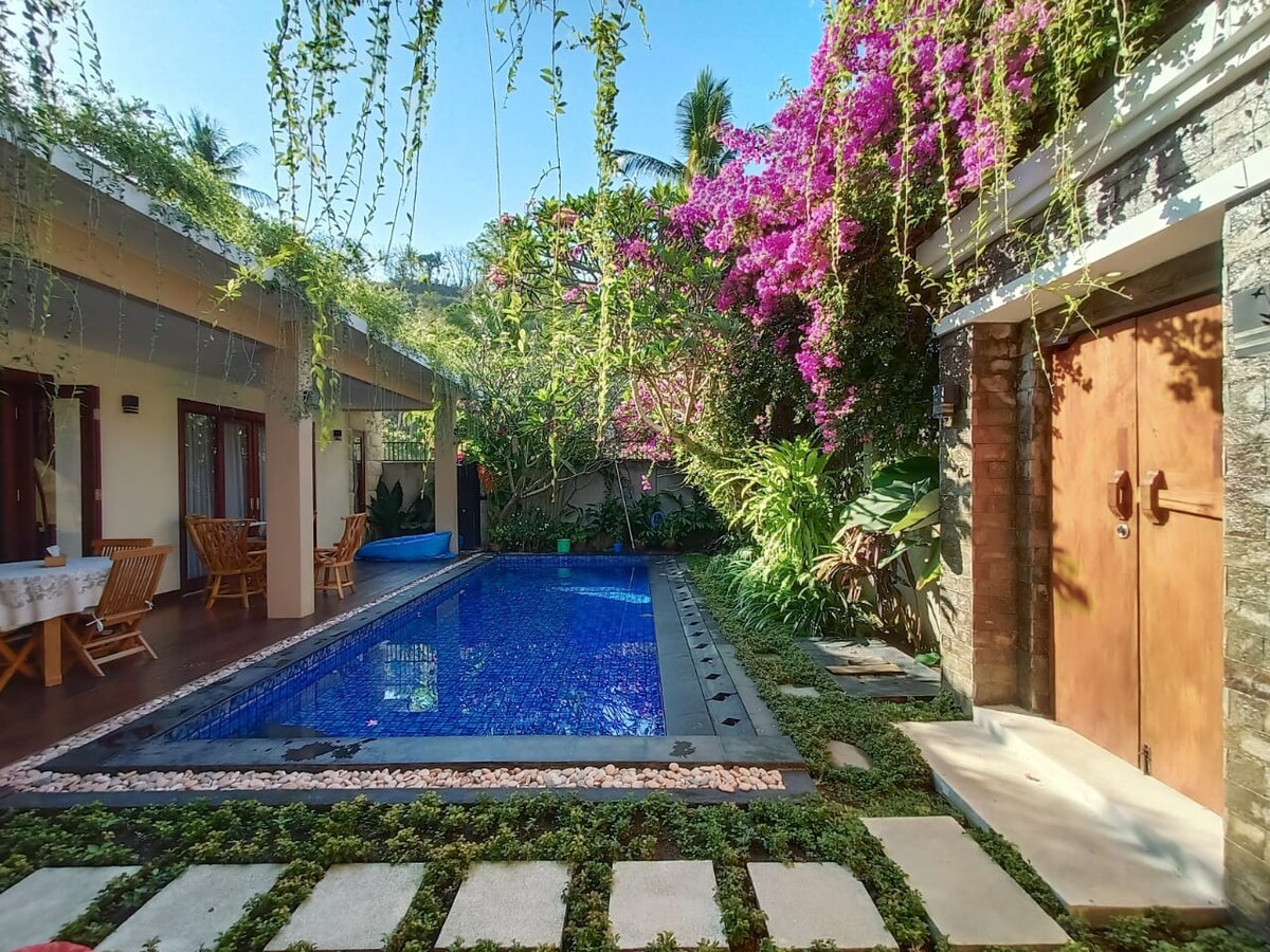 A serene outdoor area features a vibrant pool surrounded by lush greenery and flowering vines. Stone pathways lead to the pool, while wooden furniture is positioned nearby, offering a tranquil space for relaxation among nature.