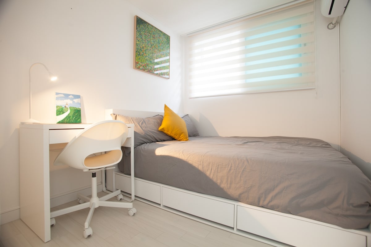 A simple bedroom features a spacious bed dressed in light gray bedding, complemented by a bright yellow accent pillow. A small desk with a chair is positioned nearby, illuminated by a desk lamp. Natural light filters through a window with horizontal blinds.