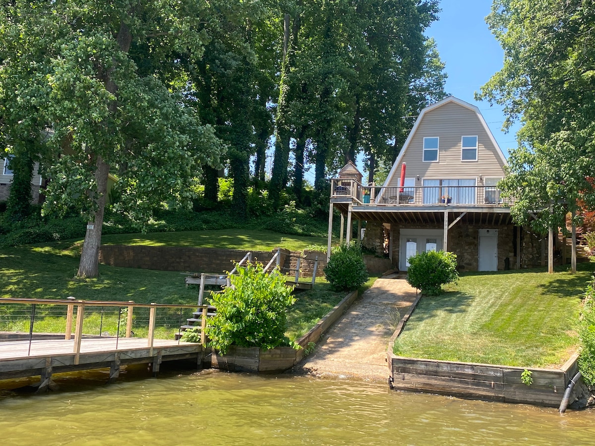 The exterior of the house is visible from the water, showcasing a two-story structure surrounded by lush green trees. A private dock extends into the lake, while a sloped path leads to the entrance of the home. A large deck overlooks the water.