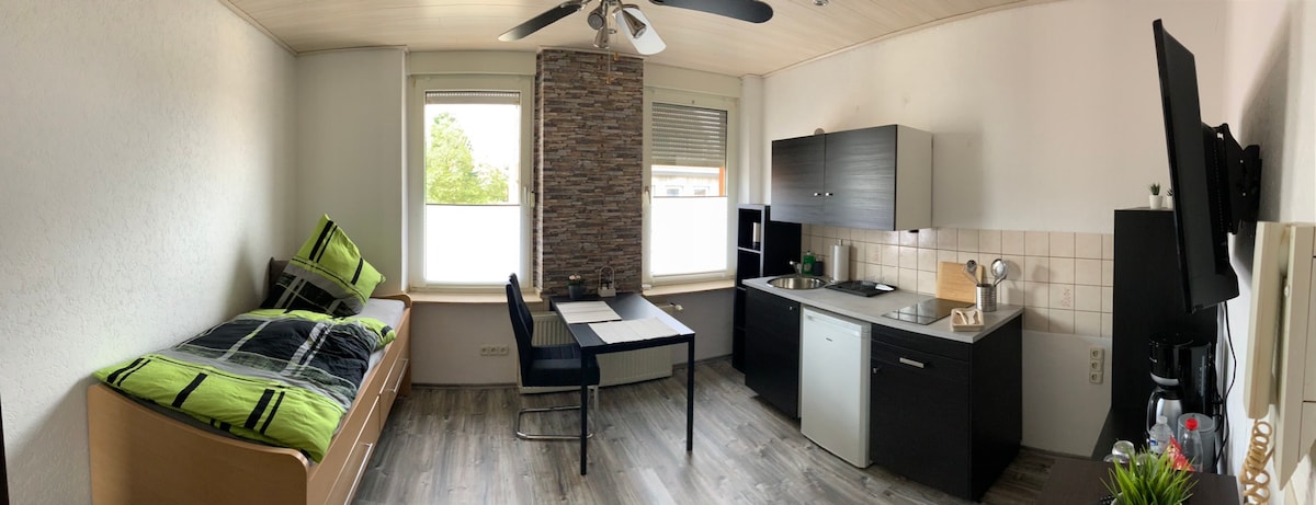 A modern room features a single bed with a green and black blanket, a dining table with two chairs, and a kitchenette equipped with essential appliances. Neutral walls complement the wooden flooring and a stone accent wall adds texture. Natural light enters through two windows.