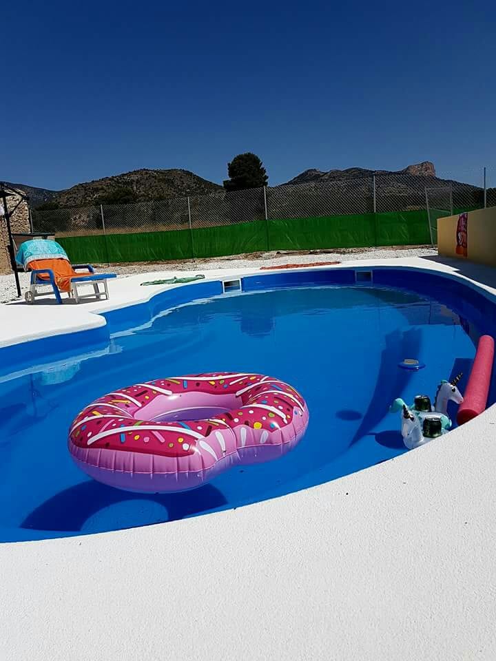A bright outdoor pool area is featured, with clear blue water reflecting the sky. A large pink donut-shaped float rests in the water. Colorful towels and lounge chairs are visible around the pool, with mountains providing a scenic backdrop.