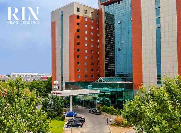 The exterior of the RIN Grand Residence is shown, featuring a modern multi-story structure with a mix of glass and brick facades. Lush greenery surrounds the entrance, where vehicles are parked under a covered drop-off area. A clear blue sky provides a backdrop.