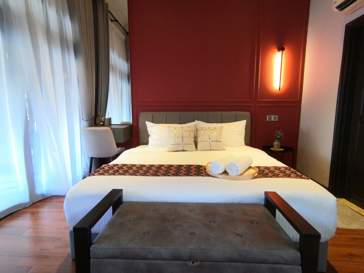 A well-arranged studio room presents a spacious bed adorned with a patterned runner. A bench sits at the foot of the bed, while light filters through the floor-to-ceiling curtains. Subtle accent lighting enhances the warm décor, creating a cozy atmosphere.