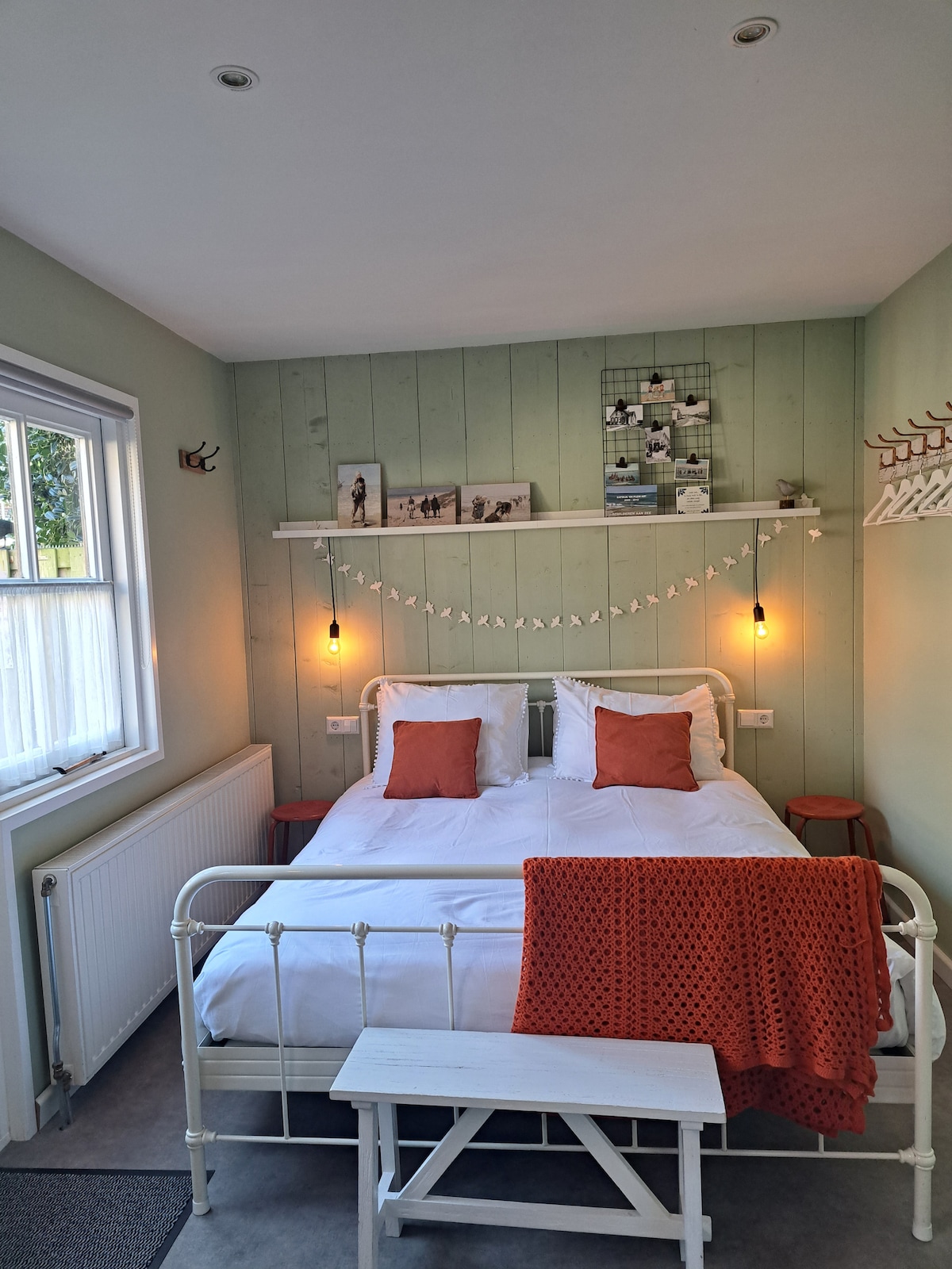 A cozy sleeping area is depicted with a white, metal bed frame holding two upright pillows and a warm blanket draped over the foot. Soft green walls provide a calming backdrop, complemented by decorative elements like wall art and indoor lighting. Natural light enters through a window on one side.