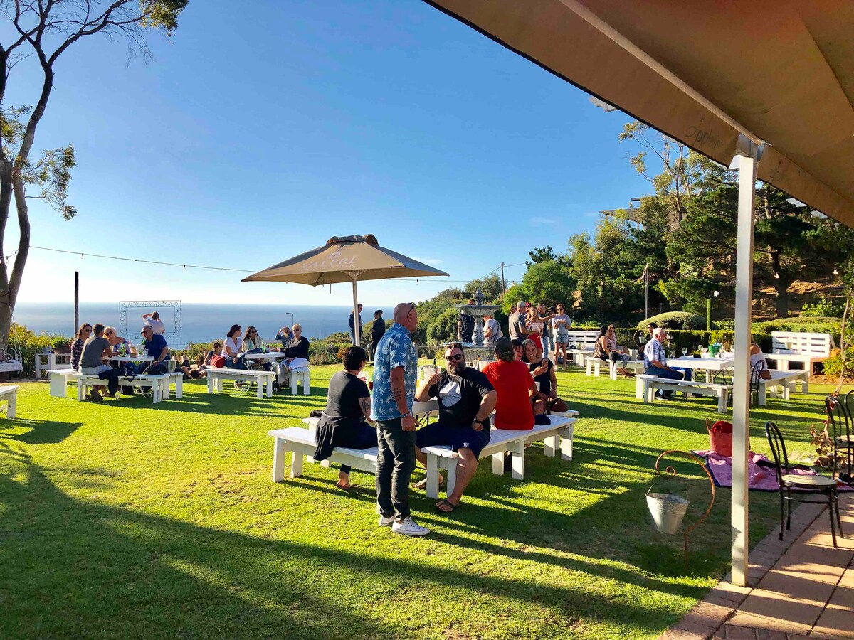 A lively outdoor seating area is showcased, featuring numerous tables and umbrellas. Guests are seen mingling on a green lawn with a view of the sea in the background. A variety of trees provide shade and enhance the relaxing atmosphere.