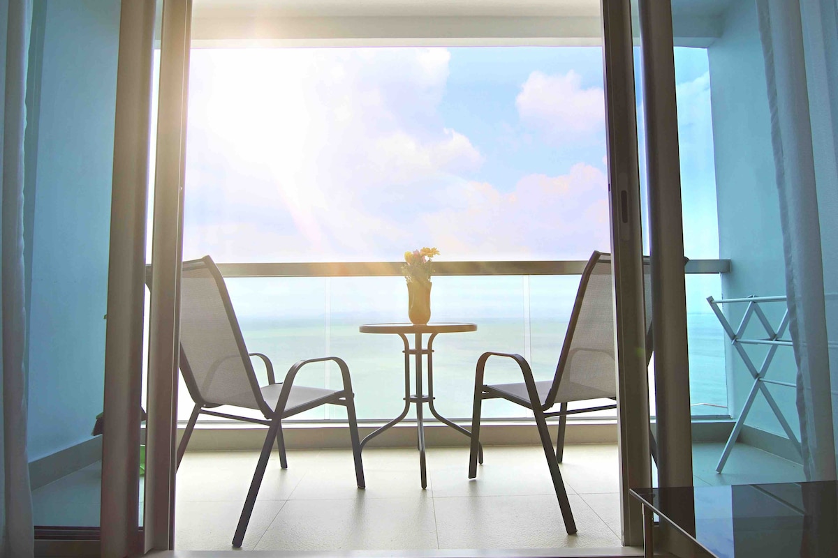 A balcony area features two chairs and a small table, positioned to overlook the ocean. Bright light filters in, creating a serene atmosphere. The glass railing provides unobstructed views, while a potted plant adds a touch of greenery to the space.