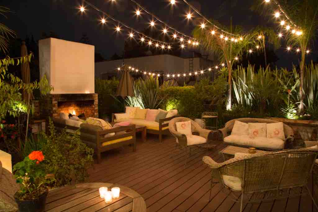 An outdoor roof deck is lit by charming overhead string lights, creating a cozy ambiance. Comfortable seating is arranged around a warm fireplace, surrounded by lush greenery and vibrant flowers, inviting relaxation under the night sky.