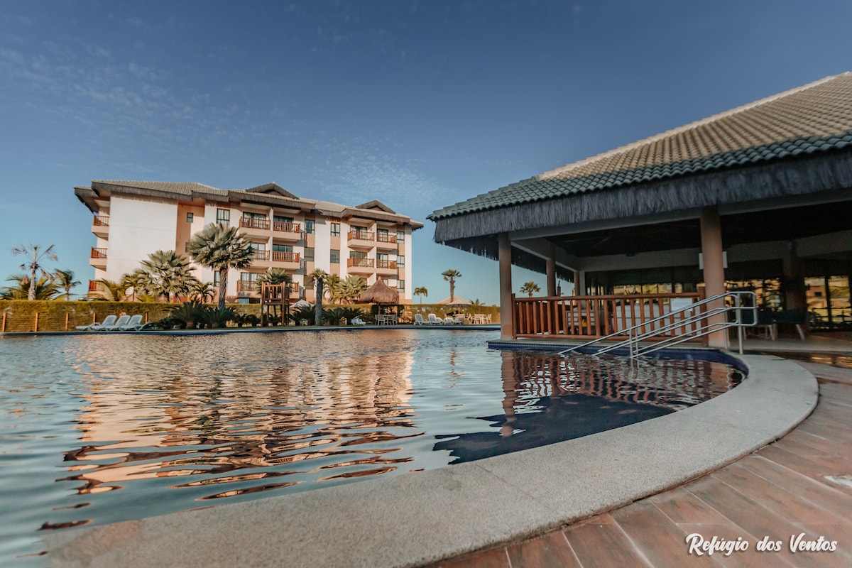 A tranquil pool area is highlighted, with a spacious building in the background. The clear water reflects the sky, and lounge chairs line the pool's edge, surrounded by palm trees and outdoor seating under a thatched roof.