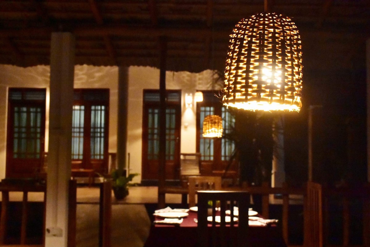 The dining area is illuminated by woven pendant lights, casting a warm glow on the dark wooden furniture. Large windows with wooden frames lead to a view of the outdoor space, while the soft lighting enhances a calm ambiance appropriate for evening meals.