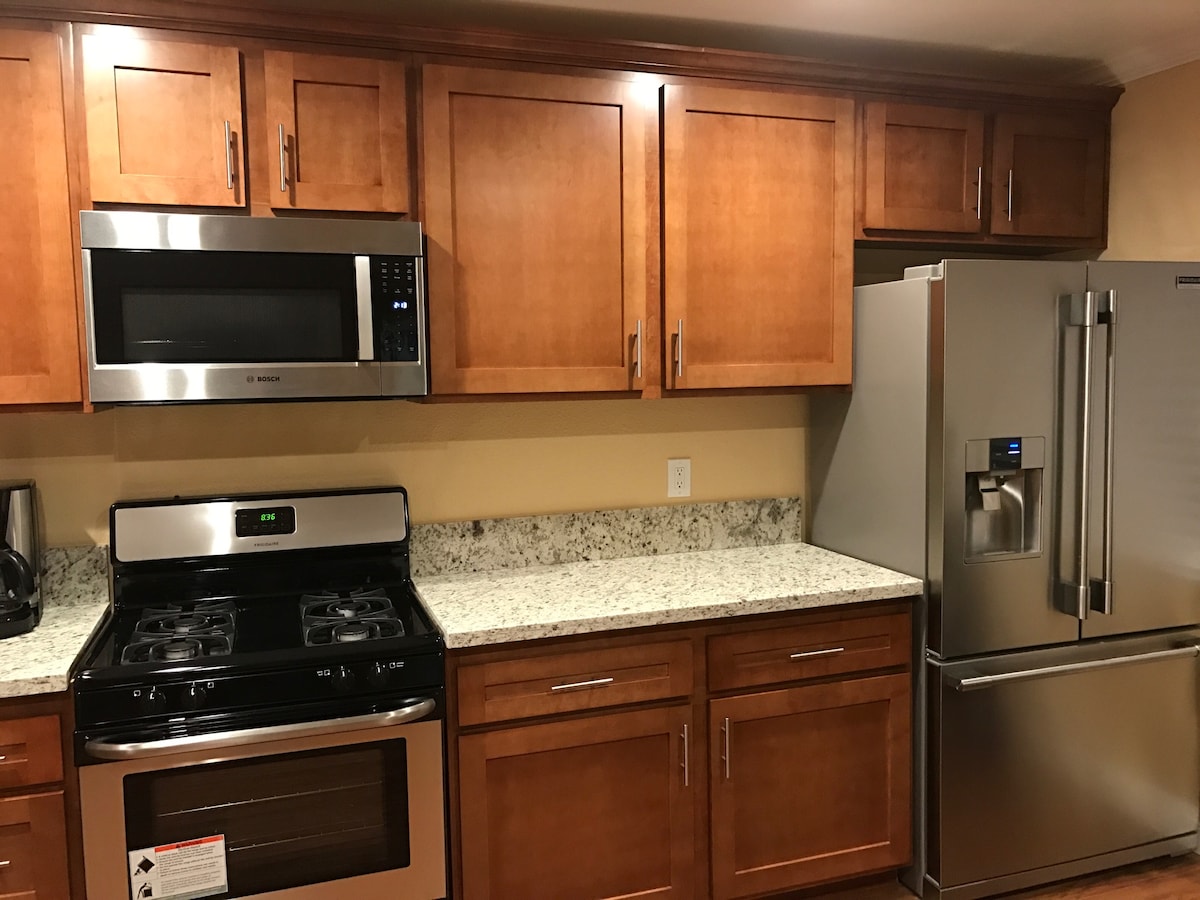 The kitchen features wooden cabinetry with a rich finish, stainless steel appliances, and a granite countertop. An oven with a gas range, a microwave, and a large refrigerator are prominently displayed. The warm lighting enhances the functional layout, providing an inviting cooking environment.