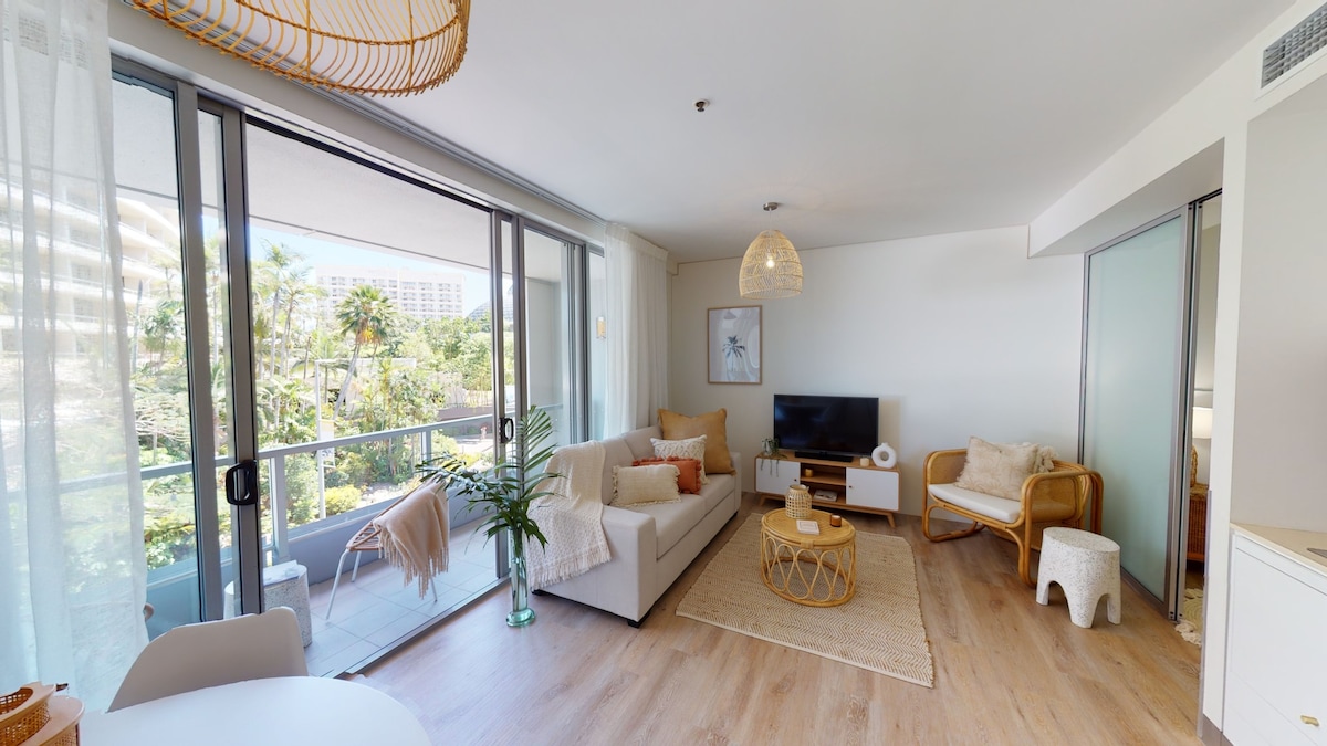 A bright and airy living area features large glass doors leading to a private balcony. A comfortable sofa is positioned beside a flat-screen TV, while a woven pendant light adds texture to the space. A small dining table sits nearby, completing the open layout.