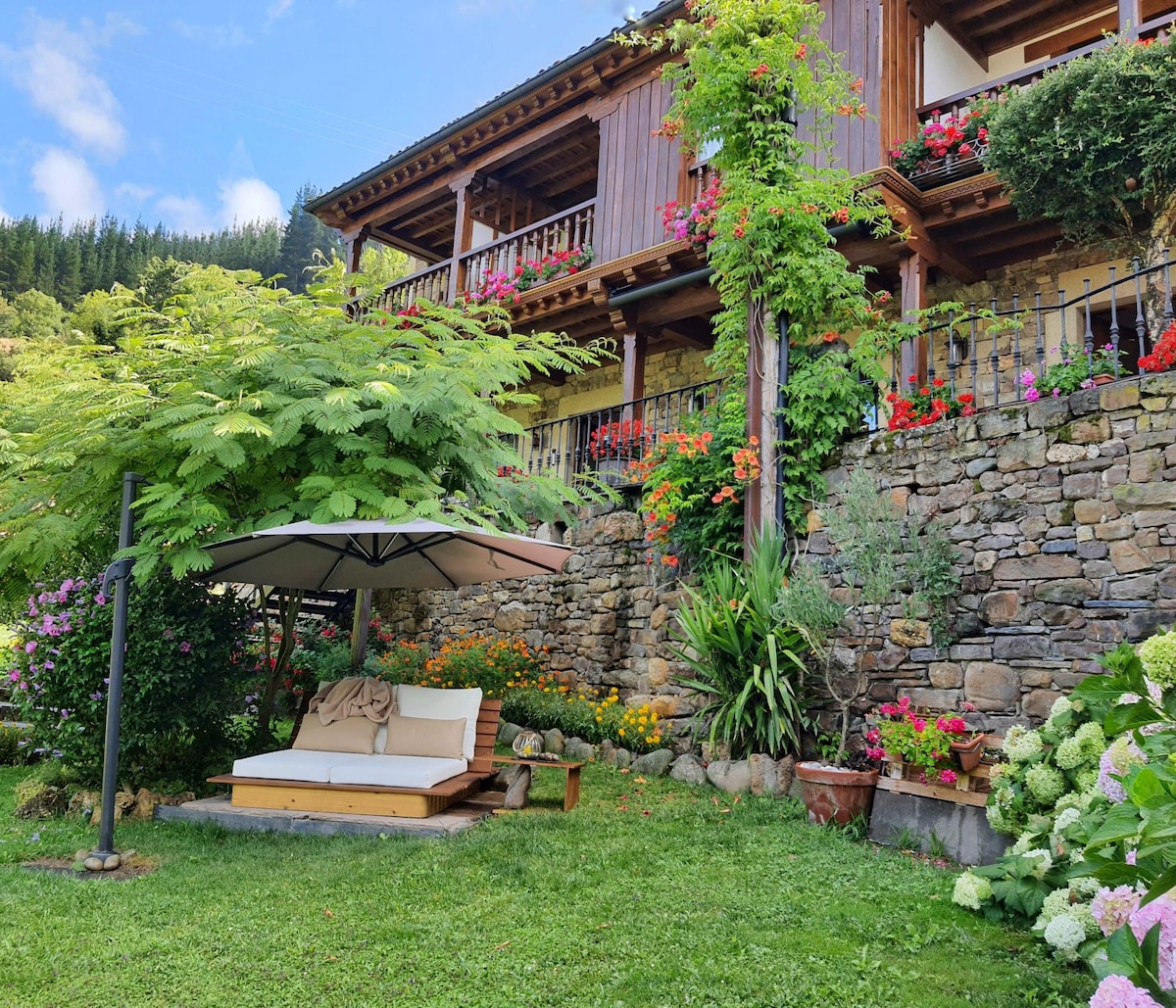 A tranquil garden area features a wooden gazebo with a comfortable lounge seating, surrounded by vibrant flowers and lush greenery. The stone wall adds a rustic charm, while the upper balcony is adorned with colorful flower pots, enhancing the inviting outdoor space.