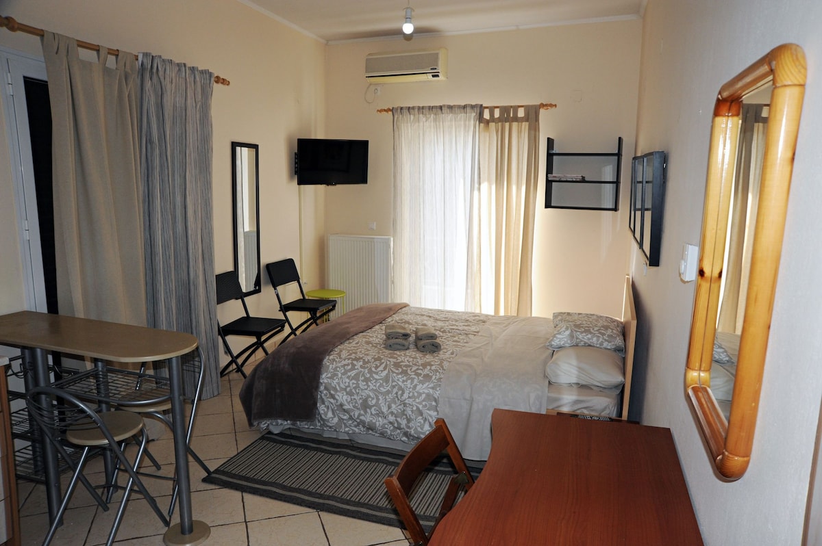 A bright room features a spacious bed dressed in neutral linens, accompanied by decorative pillows. A wooden desk and chair are present, along with a table and two folding chairs. Natural light filters through sheer curtains, providing a comfortable atmosphere.