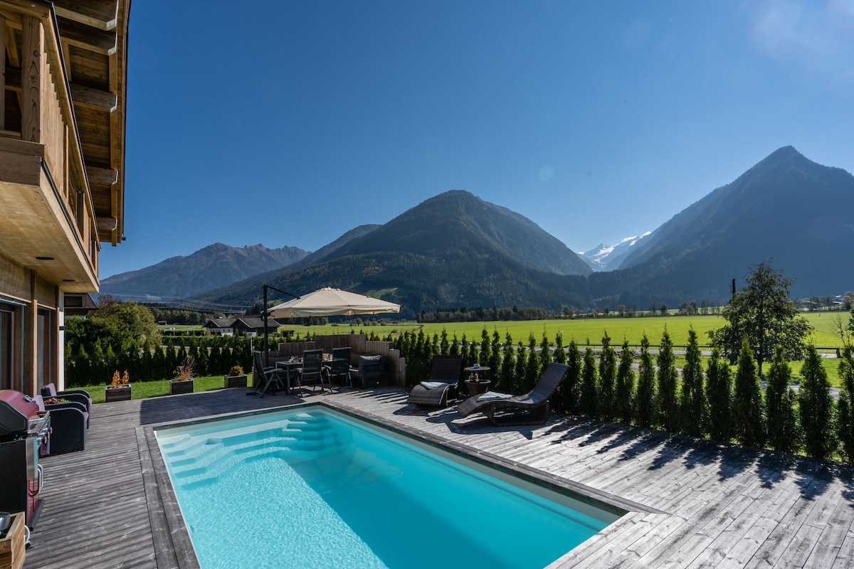 A private outdoor pool is situated on a wooden deck, surrounded by lounge chairs and an umbrella. The backdrop features expansive green fields and majestic mountains under a clear blue sky, creating a serene landscape for relaxation.