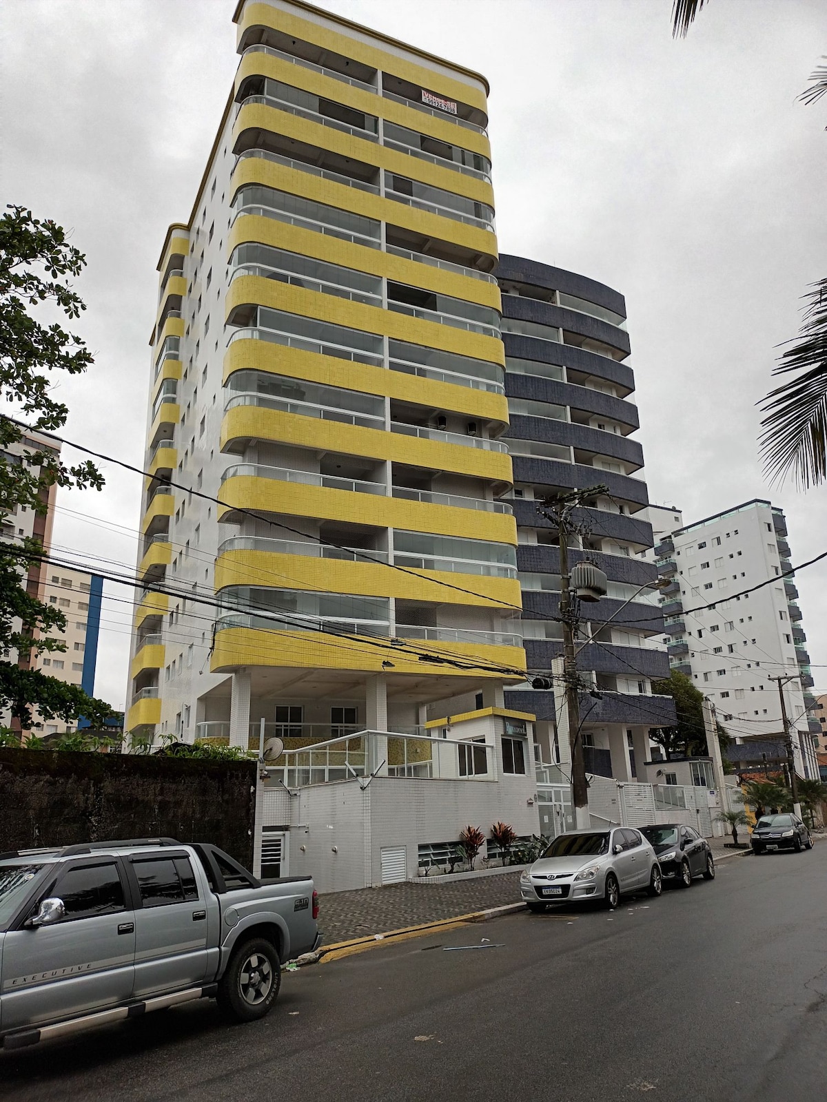 The exterior of a multi-story condominium building is shown, characterized by a yellow and gray façade. Balconies are visible on each level, while vehicles are parked along the street. The cloudy sky adds a neutral backdrop to the building's colorful design.