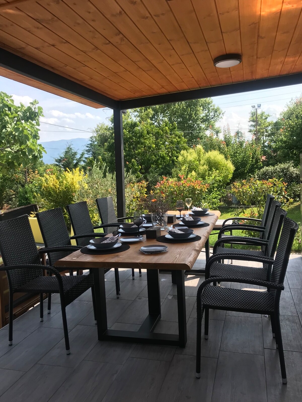 A spacious outdoor dining area is presented, featuring a large wooden table set with plates and silverware. Black wicker chairs surround the table under a wooden roof. Lush greenery and colorful plants are visible in the background, enhancing the serene ambiance.
