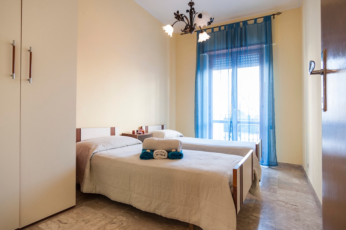 A bright bedroom features two comfortable beds, neatly made with light bedding. Natural light enters through a large window draped with sheer blue curtains. A spacious wardrobe is positioned against one wall, contributing to the room's airy feel.