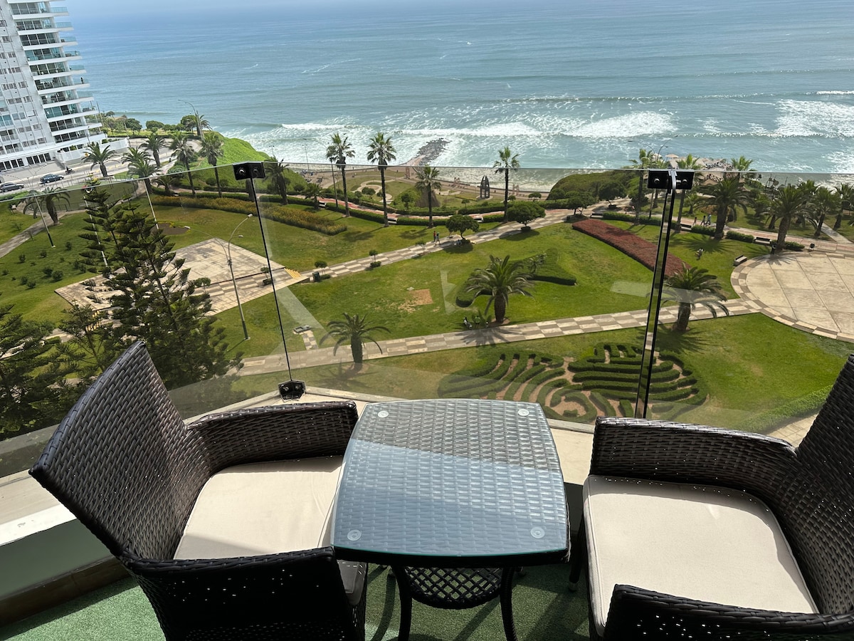 A modern terrace features two wicker chairs and a glass table, offering views of the expansive green park below, framed by palm trees. The ocean waves create a soothing backdrop, enhancing the inviting outdoor space.