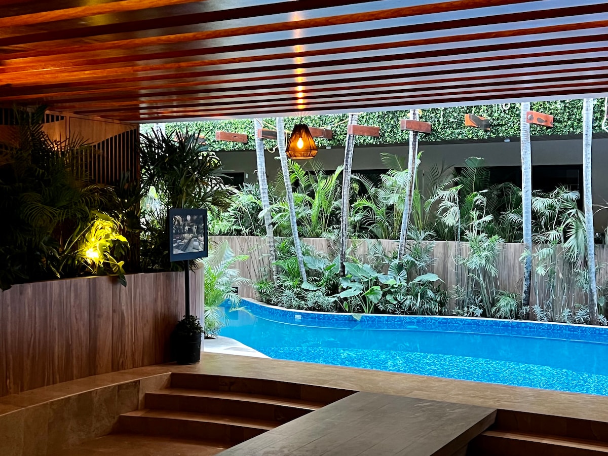 A ground-level pool area is surrounded by lush greenery and palm trees, creating a serene atmosphere. Soft lighting accentuates the wooden features, while a small outdoor seating space offers a view of the inviting blue water.