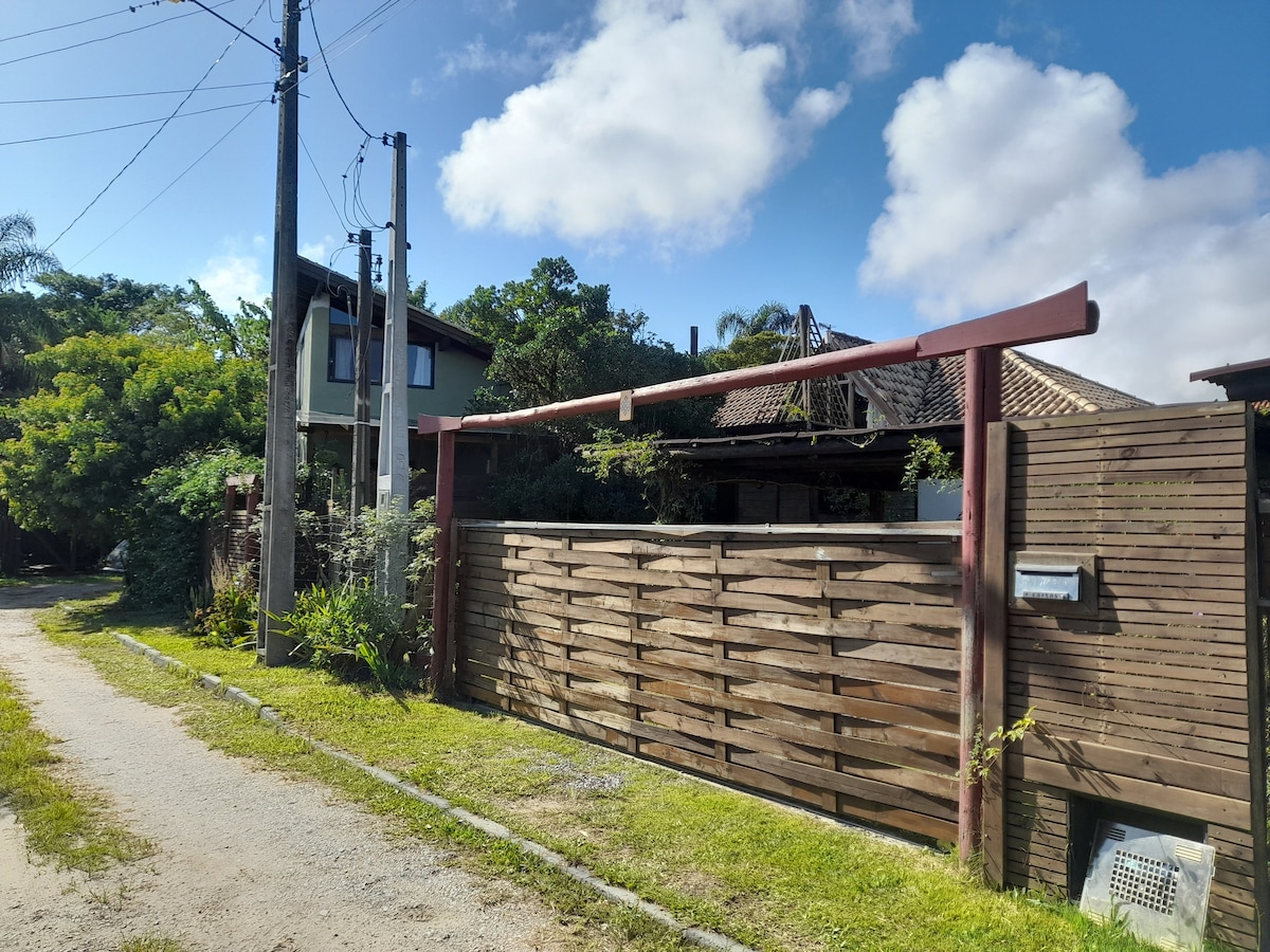 A wooden gate marked by natural greenery opens to a private path leading to the house. The structure is framed by trees, providing a quiet and secluded atmosphere. Power lines and nearby utility poles are visible along the gravel road.