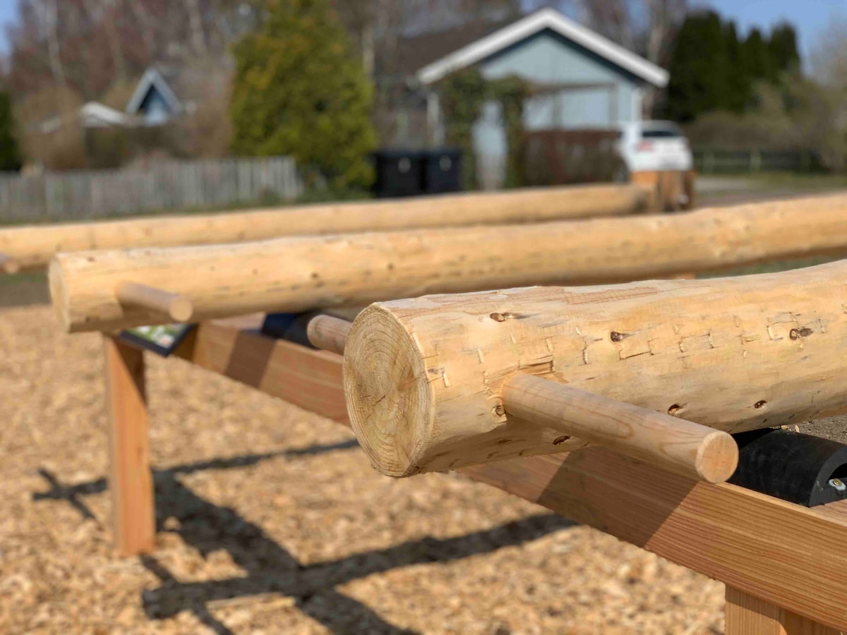 A wooden outdoor gym features parallel bars made of smooth logs, positioned on a bed of wood chips. A nearby residential setting is visible in the background, with a blue house and parked vehicle.