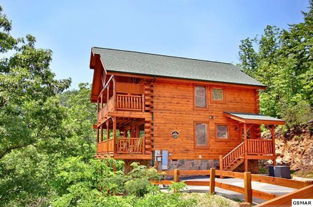 A rustic log cabin is set amidst lush trees, featuring a multi-level structure with covered porches and a pitched roof. The exterior wood finish blends harmoniously with the natural surroundings, while a wooden fence defines the property's boundary.
