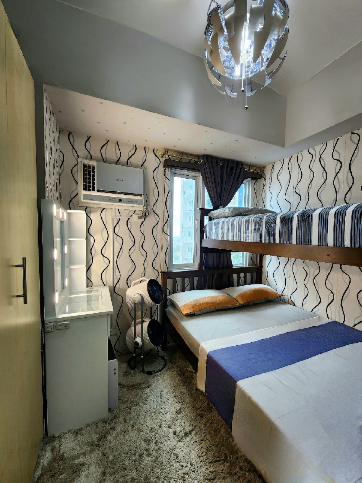 The bedroom features a layout with a single bed and a bunk bed adorned with layered bedding. A modern chandelier hangs above, with windows allowing natural light to fill the space. An air conditioning unit is positioned near the ceiling, and a vanity table is visible beside a soft carpet.
