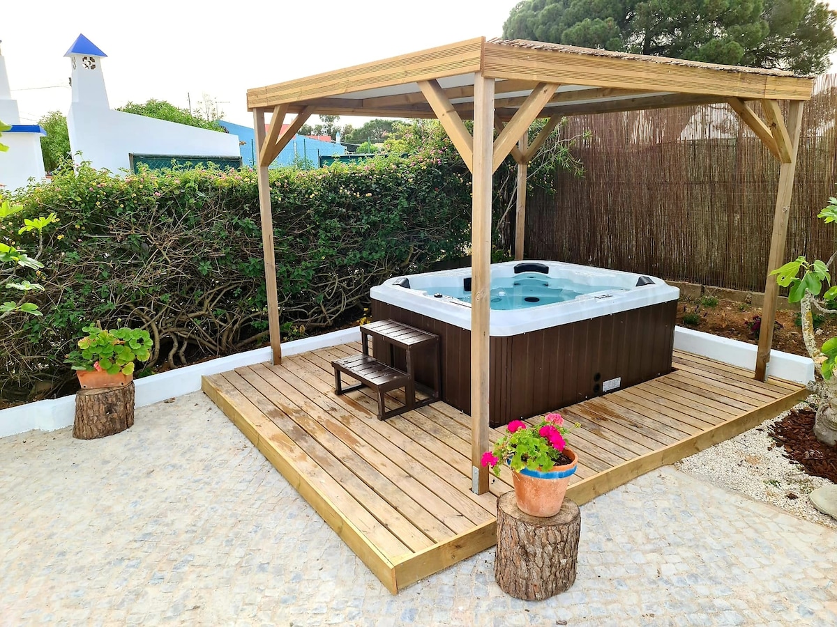 A wooden deck featuring a private jacuzzi is framed by lush vegetation. Planters with vibrant flowers add color to the setting. The area is enclosed, promoting a peaceful environment for relaxation.