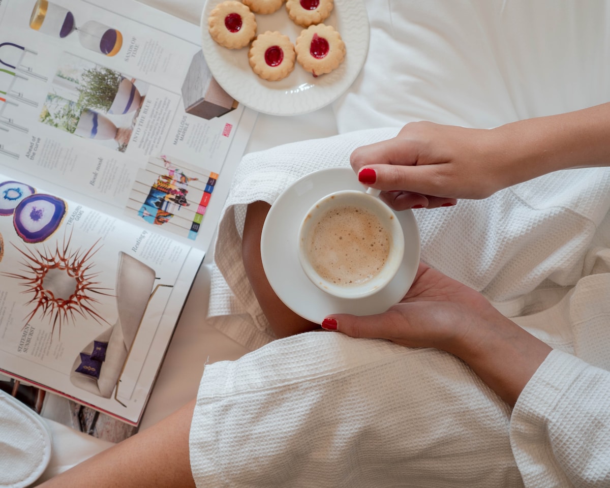 A hand holds a cup of coffee, resting on a small white saucer. Nearby, a magazine is opened, featuring a colorful layout. Fresh cookies are arranged on a plate, with a bedspread serving as the background. Light colors and a relaxed atmosphere are present.