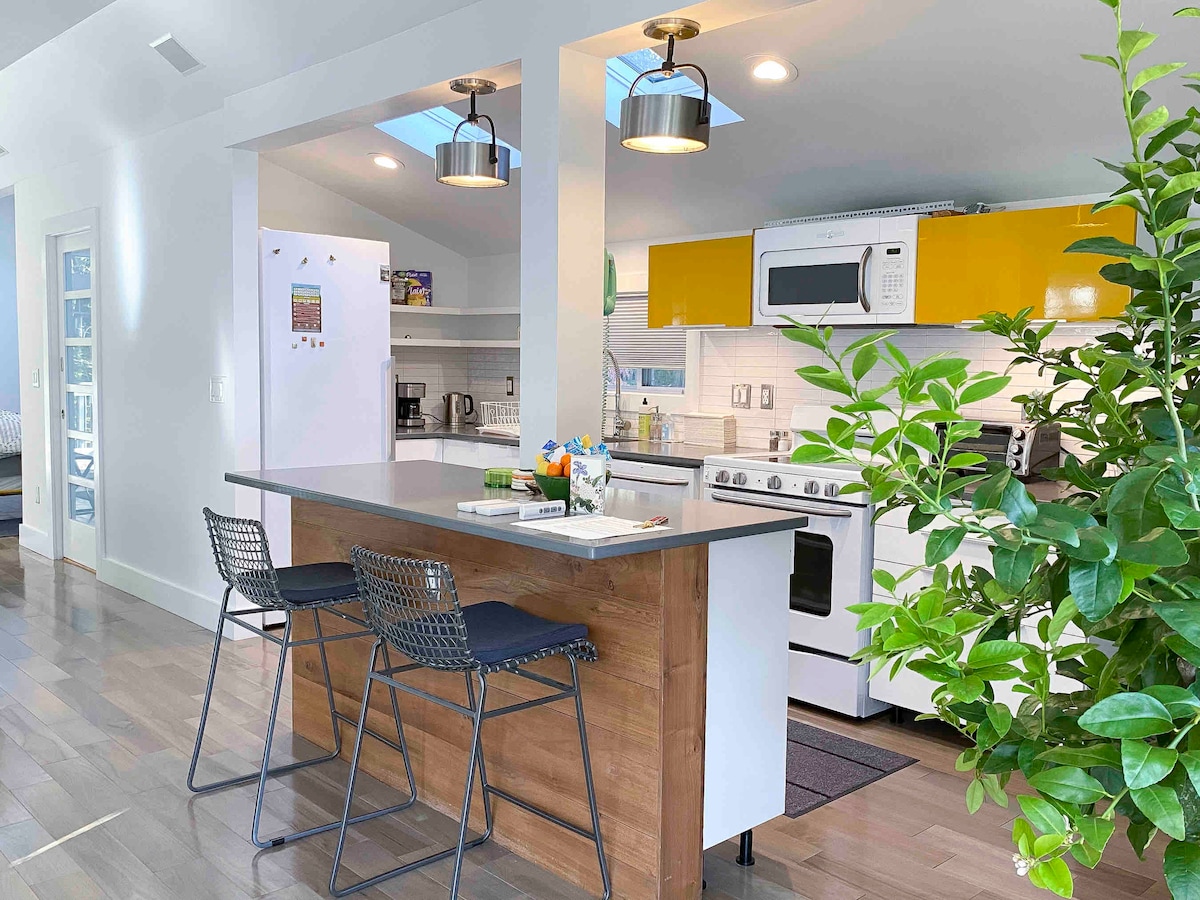 A modern kitchen area is highlighted by a central island with two textured bar stools. Bright yellow cabinetry complements the stainless steel appliances. Natural light filters in through skylights, and a green plant adds a touch of nature to the space.
