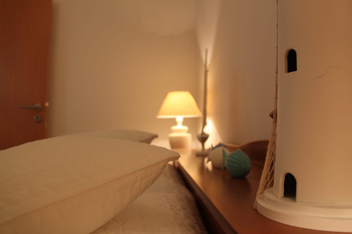 A serene bedroom scene features a soft white quilt laid over a spacious bed. Pillows are arranged neatly, and a warm lamp casts a gentle glow. A charming decorative lighthouse adds a nautical touch, enhancing the cozy environment.