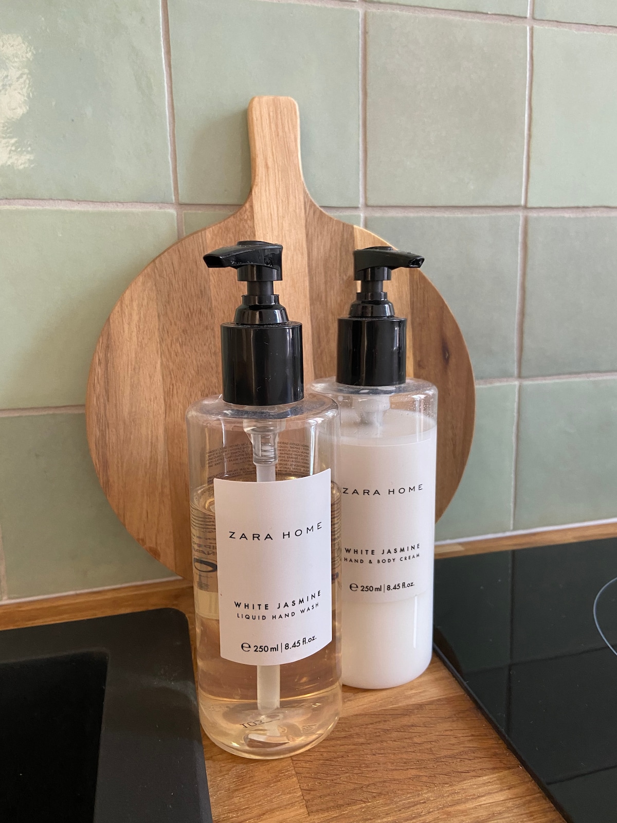 A wooden cutting board supports two bottles of liquid soap and lotion. Both bottles feature a minimalistic design with black pumps and white labels. The background displays a tiled surface in soft green tones, complementing the items in the foreground.
