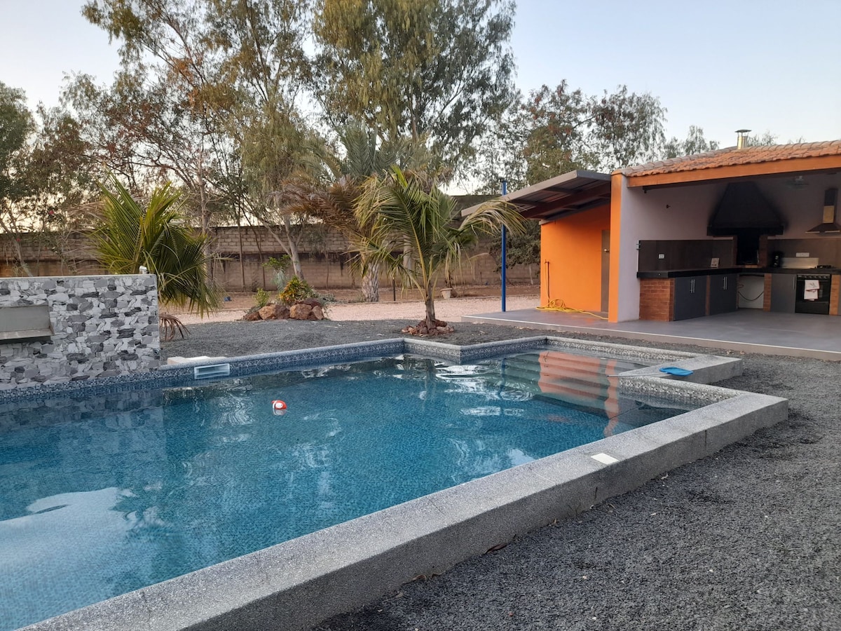 The outdoor area features a refreshing swimming pool bordered by sleek stone and surrounded by gravel landscaping. A shaded seating area with an outdoor kitchen and barbecue grill is visible in the background, set against vibrant orange walls and lush greenery.