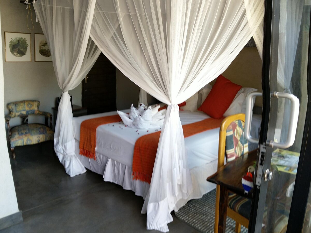 A spacious sleeping area features a large bed draped with sheer canopies, complemented by vibrant orange and white bedding. A cozy armchair is positioned nearby, adding to the room's comfort. A rustic wooden table holds decorative items, and natural light floods in through the sliding door.