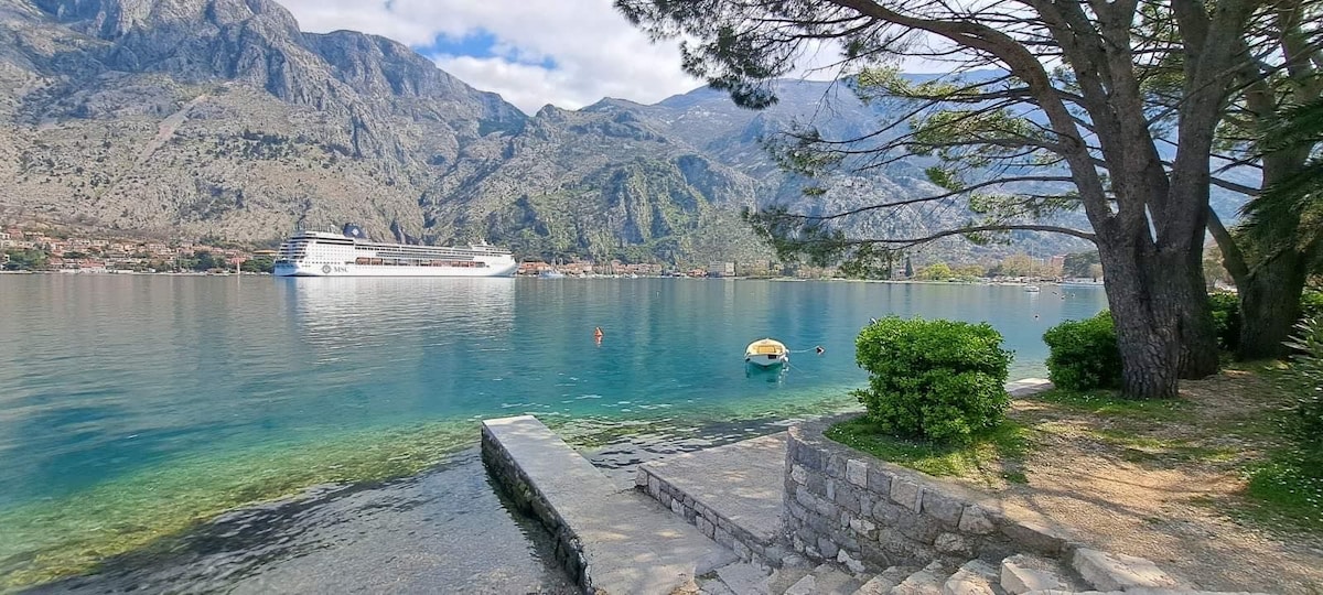 A serene waterfront scene features clear turquoise waters gently lapping against a stone path. Lush greenery and tree shadows are visible along the shore, while a small boat floats nearby. Majestic mountains rise in the background, providing a peaceful and natural setting.