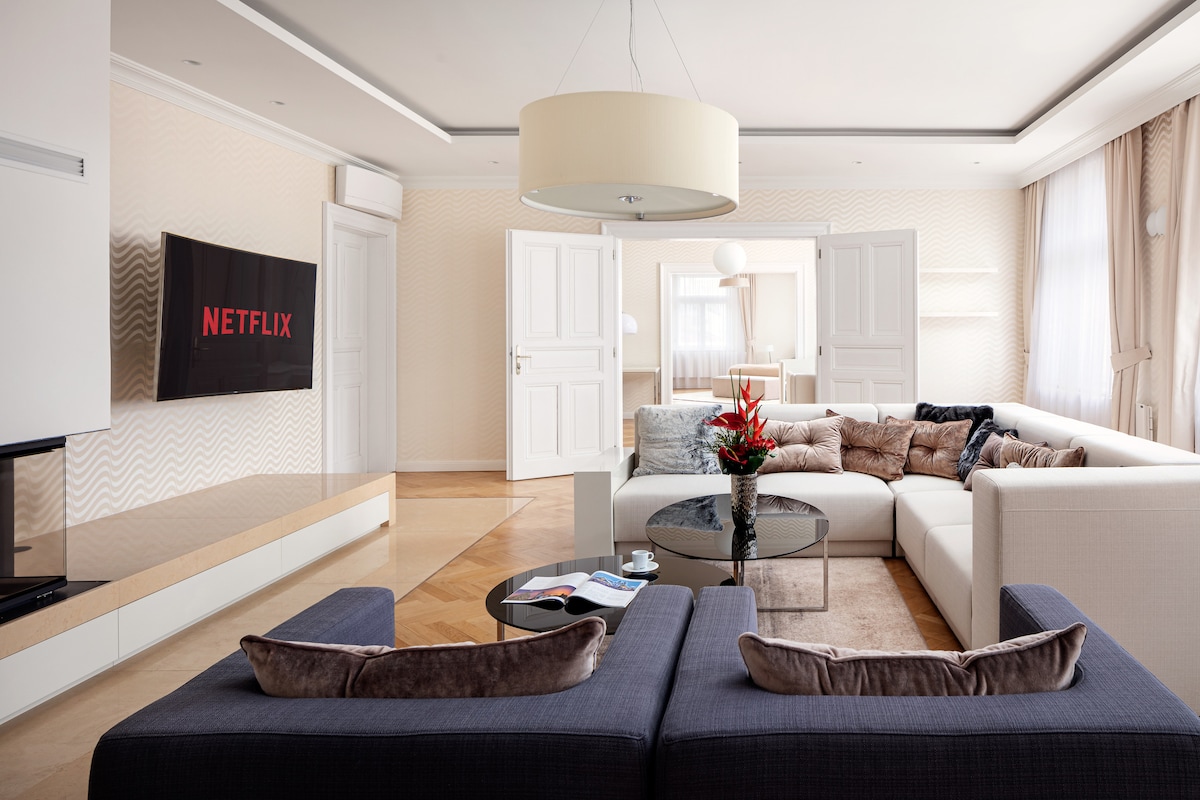 A spacious living room features a large sectional sofa facing a wall-mounted television displaying Netflix. A round glass coffee table sits in the center, surrounded by decorative plants. Natural light enters through large windows, adding warmth to the neutral-toned decor and furnishings.