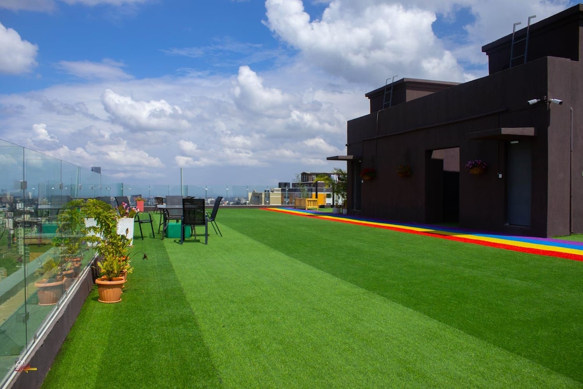 A spacious rooftop terrace features vibrant artificial grass, complemented by colorful pathways. Modern seating arrangements with chairs are visible. Potted plants add touches of greenery, while the expansive skyline can be seen in the background beneath a partly cloudy sky.