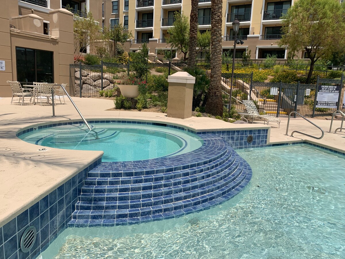 A sparkling hot tub is situated beside a community pool, featuring tiled steps that lead into the warm water. Surrounding landscaped areas include palm trees and stone accents, with seating arrangements visible in the background. Safety handrails are provided for easy access.