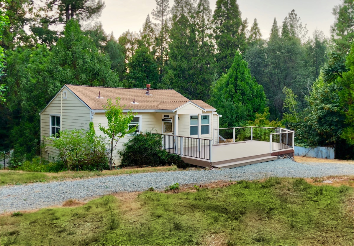 A quaint cottage sits amidst a natural setting, framed by tall trees and lush greenery. A spacious deck extends from the front, providing an inviting area for relaxation. The gravel pathway leads to the entrance, enhancing the serene atmosphere of the surrounding landscape.