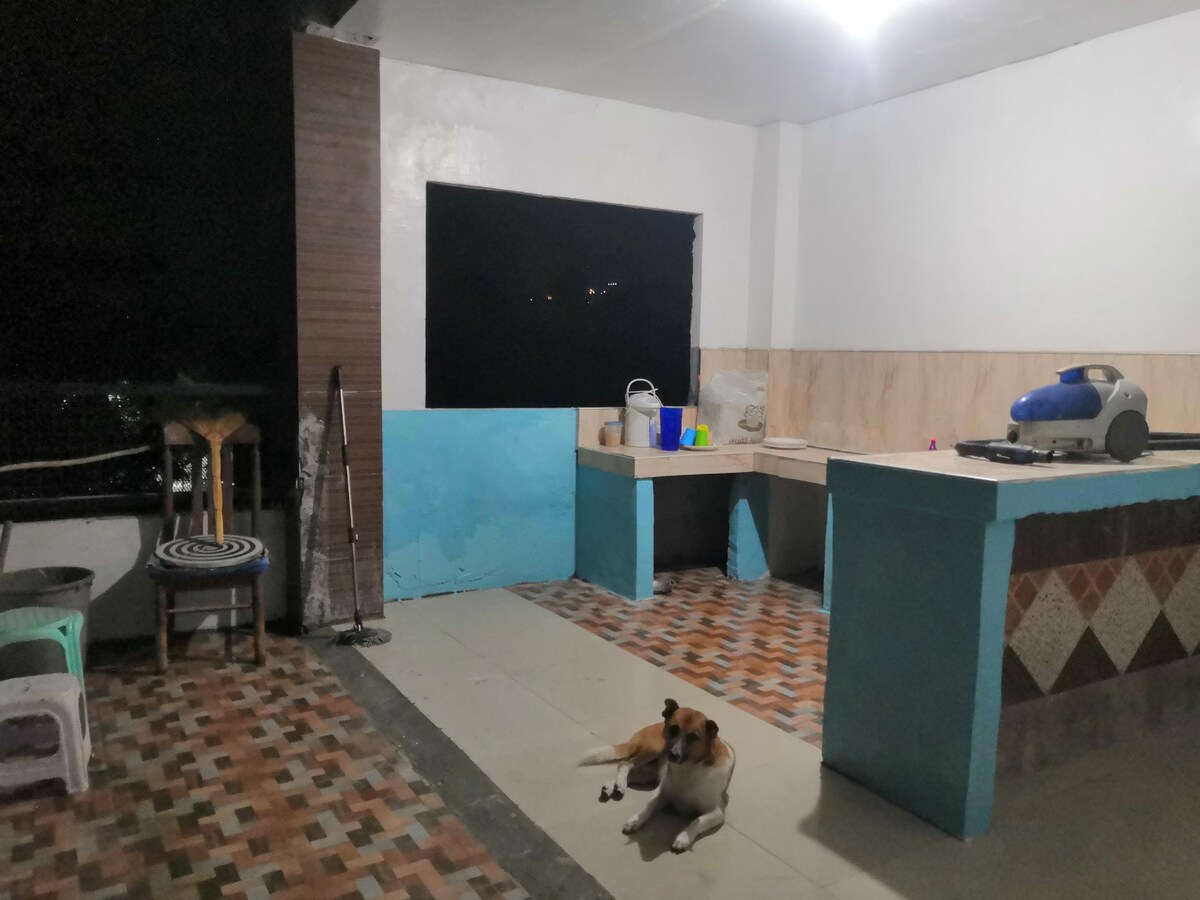A spacious outdoor terrace is illuminated by soft lighting, featuring a simple kitchenette area with a counter and a few kitchen supplies. A dog rests peacefully on the tiled floor, and seating options are available in the corner.