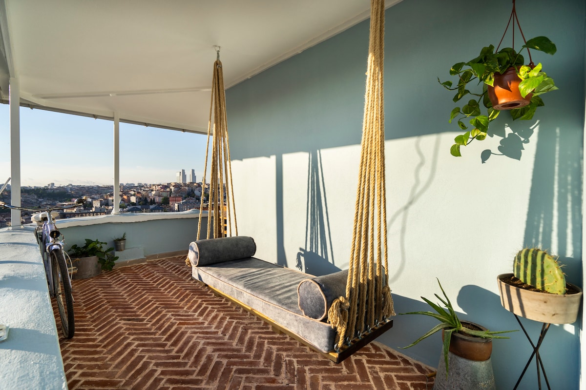 A spacious terrace features a suspended swing, draped with soft fabric, providing a relaxing spot to enjoy city views. Potted plants surround the area, including a hanging plant and a cactus, while a bicycle leans against the wall, offering charm to the outdoor space.